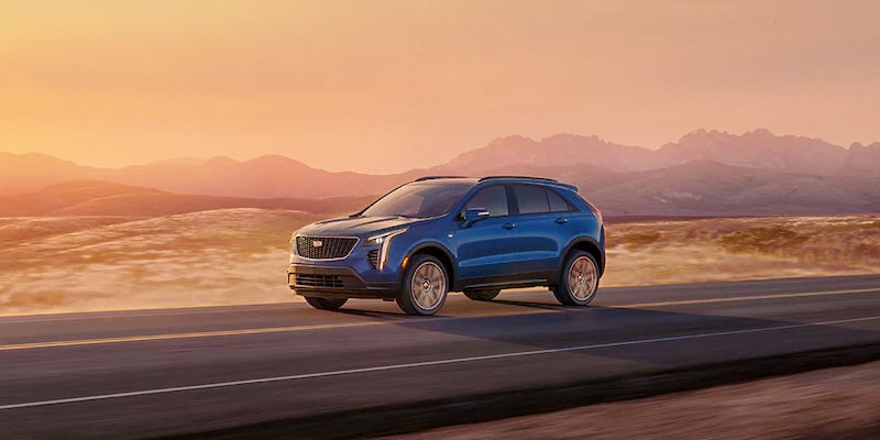 Blue Cadillac XT4 driving on an open road through a desert landscape at sunset, with mountain silhouettes in the background.