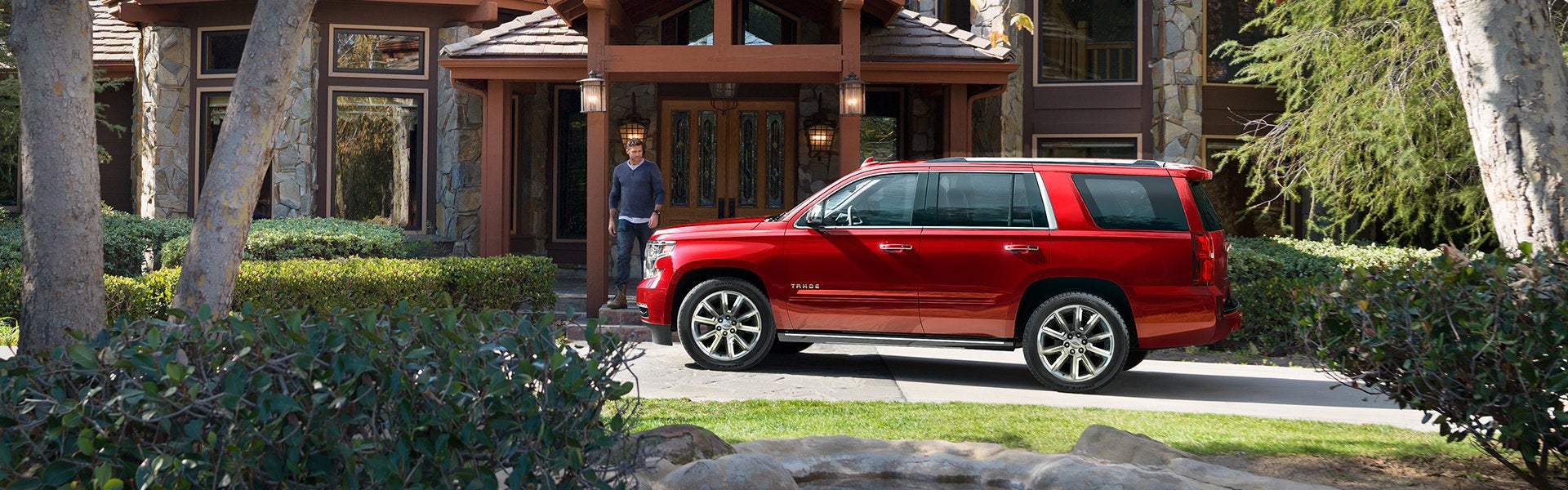 A red Chevrolet Tahoe SUV parked in the driveway of a rustic stone-and-wood house, with a man stepping out of the front entrance and landscaped shrubs and trees framing the scene.