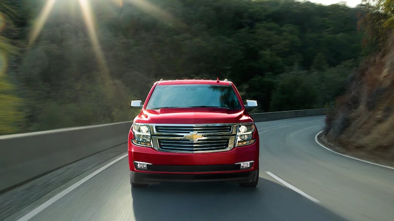 A red Chevrolet Tahoe driving on a curving mountain road during daylight, surrounded by trees and guardrails.