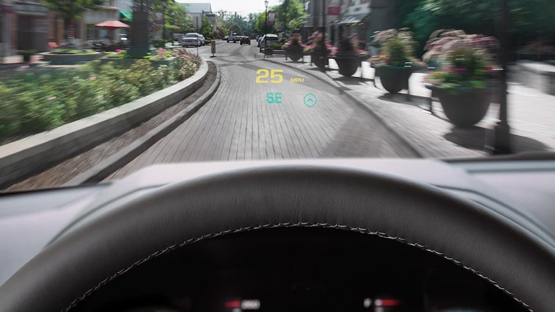 Driver’s perspective showing a head-up display on the windshield, indicating a speed of 25 MPH and southeast direction while driving through a landscaped urban area.