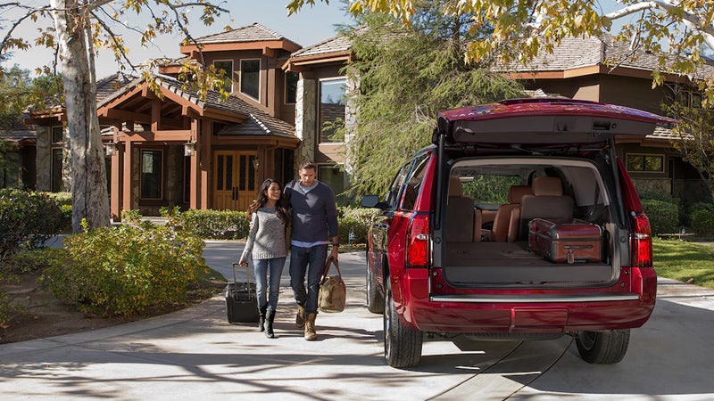 "A couple walking toward a red Chevrolet SUV parked in the driveway of a large home, with the rear liftgate open and suitcases loaded inside, suggesting preparation for a trip.