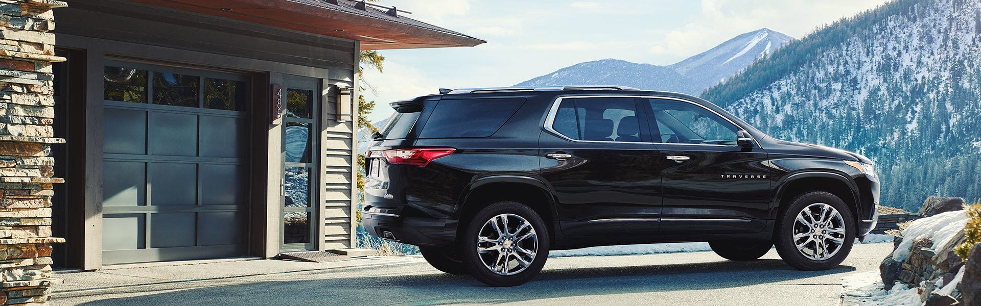 A black Chevrolet Traverse SUV parked in front of a modern cabin-style garage with snow-covered mountains and pine trees in the background.