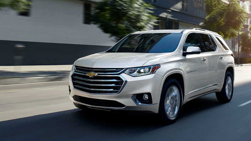 A white Chevrolet Traverse SUV driving on a city street in daylight, with motion blur suggesting speed and urban surroundings.