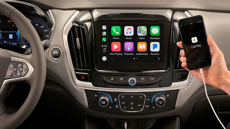 Interior view of a Chevrolet SUV dashboard with Apple CarPlay displayed on the infotainment screen, as a hand connects an iPhone.