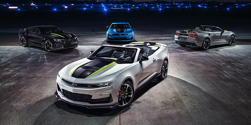 Four Chevrolet Camaro convertibles parked under night lights, featuring various models in black, white, blue, and silver with sporty accents.