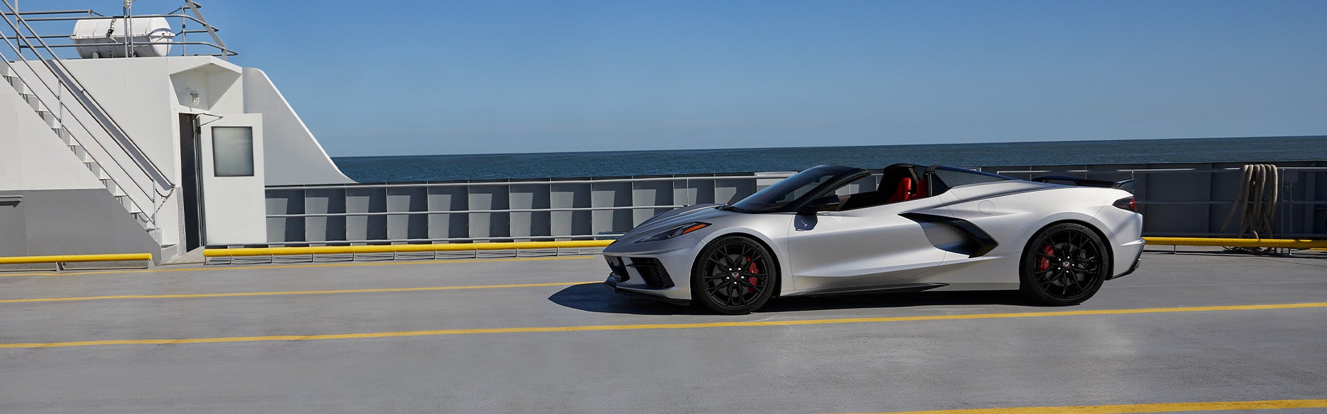 A silver convertible sports car with black wheels and red brake calipers parked on a ferry deck with the ocean and clear blue sky in the background.