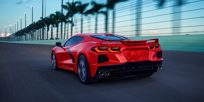 A red Chevrolet Corvette driving on a racetrack-like road lined with palm trees during sunset.