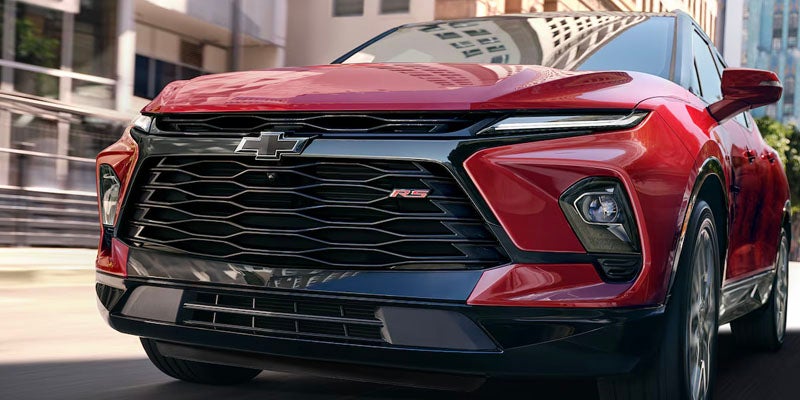 Close-up front view of a red 2025 Chevrolet Blazer RS driving through a city street, showcasing its bold black grille and sleek headlights.