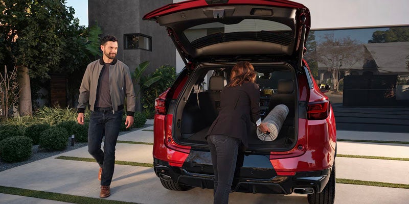 Rear view of a red 2025 Chevrolet Blazer with the liftgate open as a woman loads a rolled-up rug while a man walks nearby.
